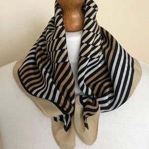May include: A square scarf featuring a geometric pattern of black, white, and tan diagonal stripes. The design includes sections of alternating lines and triangular shapes, finished with a solid tan border along the edges of the fabric.