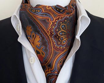 May include: A patterned ascot in shades of brown, gold, red, and blue. The silk cravat features an intricate paisley design and is worn with a white collared shirt and a black suit jacket. The cravat is a formal accessory.