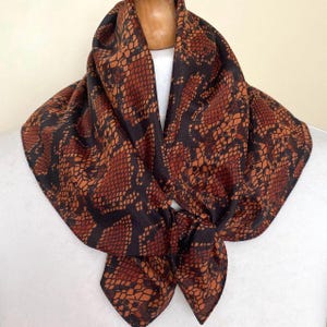 Puede incluir: Un pañuelo cuadrado con un estampado de piel de serpiente en tonos marrones y negros. El pañuelo está anudado en la parte delantera, mostrando el intrincado diseño. Se muestra sobre una superficie blanca.