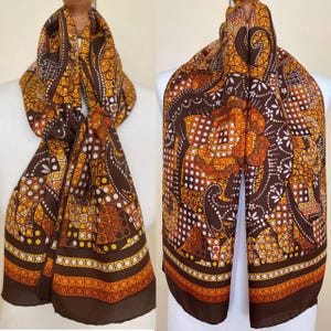May include: A brown and orange patterned scarf with a repeating geometric design. The scarf features a dark brown border and is draped over a white form. The pattern includes circles, squares, and floral elements.