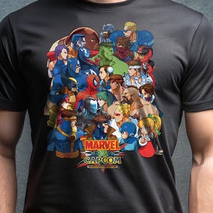 May include: Dark gray t-shirt featuring a colorful graphic of multiple Marvel and Capcom characters. The design includes the text "MARVEL vs. CAPCOM" in a stylized font. The shirt is made of a soft material.
