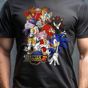 May include: Black t-shirt featuring a colorful graphic of characters from the Sonic Adventure 2 video game. The design includes Sonic, Tails, Knuckles, Amy, and Dr. Eggman. The text "SONIC ADVENTURE 2" is also visible.