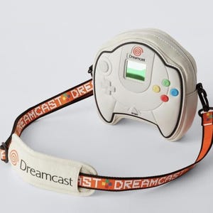 Può includere: Borsa a tracolla color crema a forma di controller Dreamcast, con pulsanti colorati e uno schermo. La borsa ha un bordo nero e una tracolla con marchio Dreamcast e un motivo arancione e nero.