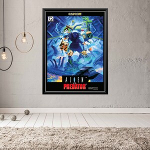 Op de afbeelding: Ingelijste poster met de artwork voor de videogame "Alien vs. Predator". De poster heeft een zwart frame en toont een levendige, actievolle scène met personages in tinten blauw, groen en oranje. De titel van het spel staat prominent in het rood.