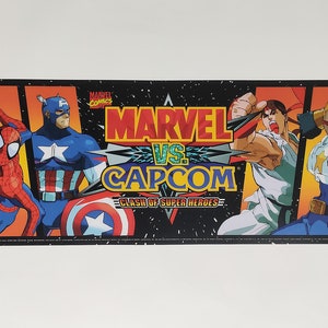 Könnte beinhalten: Ein schwarzer und orangefarbener Arcade-Schrank-Marquee mit dem Text "MARVEL VS. CAPCOM CLASH OF SUPER HEROES" und Bildern von Charakteren aus dem Spiel, darunter Wolverine, Spider-Man, Captain America, Ryu, Guile und Mega Man.