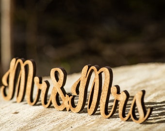 May include: Wooden cutout letters spelling "Mr & Mrs" in a cursive font. The letters are brown and are sitting on a light brown wooden surface.