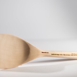 May include: A wooden spatula with a wide, flat blade and a long handle. The handle is engraved with the text "Nussbaumkronen" and "Echt statt billig, Sicher, Natur, Made in Germany".