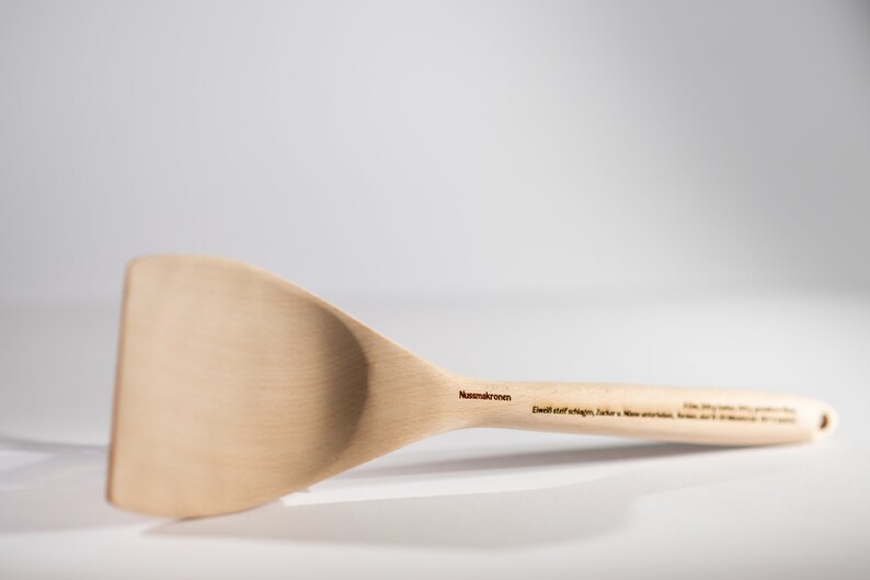 May include: A wooden spatula with a wide, flat blade and a long handle. The handle is engraved with the text "Nussbaumkronen" and "Echt statt billig, Sicher, Natur, Made in Germany".