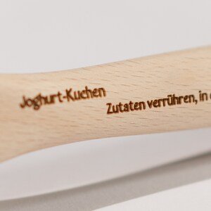 May include: A wooden spoon with the words "Yogurt-Kuchen" and "Zutaten verr&uuml;hren, in eine gefettete" engraved on it. The spoon is light brown in color.