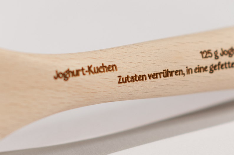 May include: A wooden spoon with the words "Yogurt-Kuchen" and "Zutaten verr&uuml;hren, in eine gefettete" engraved on it. The spoon is light brown in color.