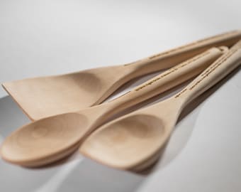 May include: Three wooden spoons, one with a rounded bowl and two with flat, wide bowls. The spoons are made of light wood and have a natural finish. The handles are engraved with text.