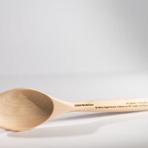May include: A wooden spoon with a light brown finish. The spoon has a rounded bowl and a long handle. The text "Einfach N&uuml;tzliche K&uuml;che" is engraved on the handle.