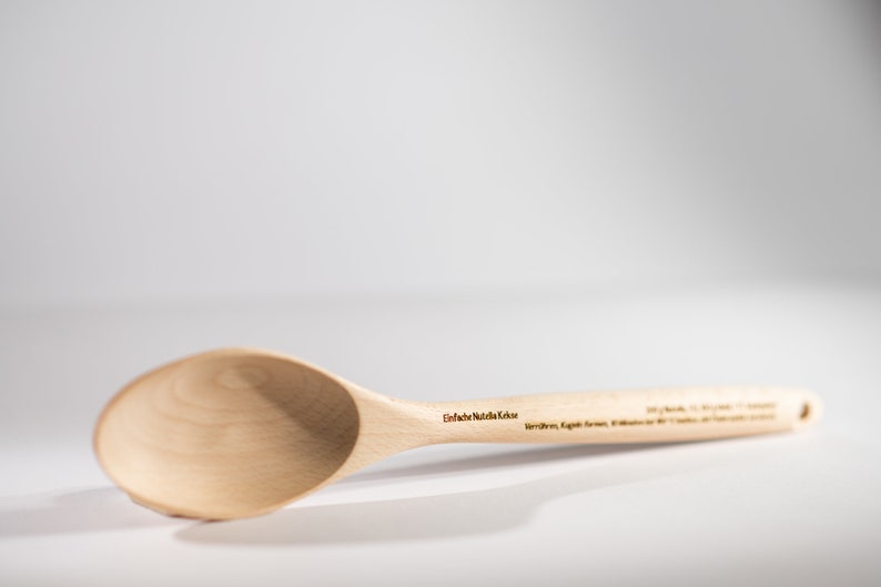 May include: A wooden spoon with a light brown finish. The spoon has a rounded bowl and a long handle. The text "Einfach N&uuml;tzliche K&uuml;che" is engraved on the handle.