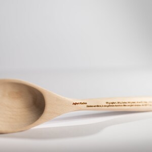May include: A wooden spoon with a rounded bowl and a long handle. The handle is engraved with the text "Joghurt L&ouml;ffel" and "Zutaten werden... in die ge&ouml;ffnete Flasche hinein..."