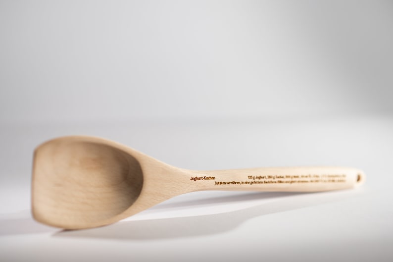 May include: A wooden spoon with a rounded bowl and a long handle. The handle is engraved with the text "Joghurt L&ouml;ffel" and "Zutaten werden... in die ge&ouml;ffnete Flasche hinein..."