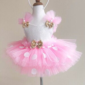 May include: A pink tulle tutu with white polka dots and a white bodice with gold bows. The tutu is worn by a mannequin and has a gold headband with pink bow and gold ears.
