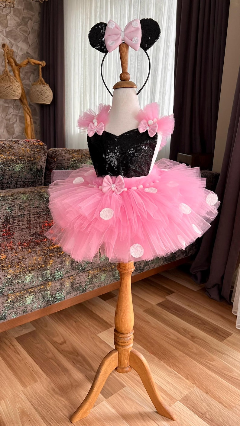 Pu&ograve; includere: Una gonna tutu in tulle rosa con pois bianchi e un corpetto nero di paillettes con fiocchi rosa. La gonna &egrave; su un manichino di legno con un cerchietto nero con orecchie rosa e un fiocco.