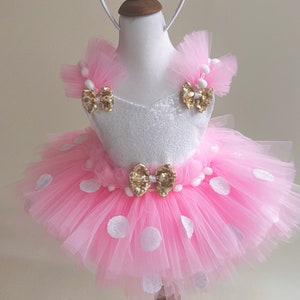 May include: A pink tulle tutu skirt with white polka dots and a white bodice with gold sequins and bows. The skirt is worn by a mannequin and has a white pom-pom trim. The mannequin is wearing a gold headband with pink bow and gold ears.