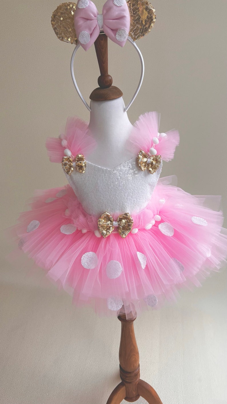 May include: A pink tulle tutu skirt with white polka dots and a white bodice with gold sequins and bows. The skirt is worn by a mannequin and has a white pom-pom trim. The mannequin is wearing a gold headband with pink bow and gold ears.