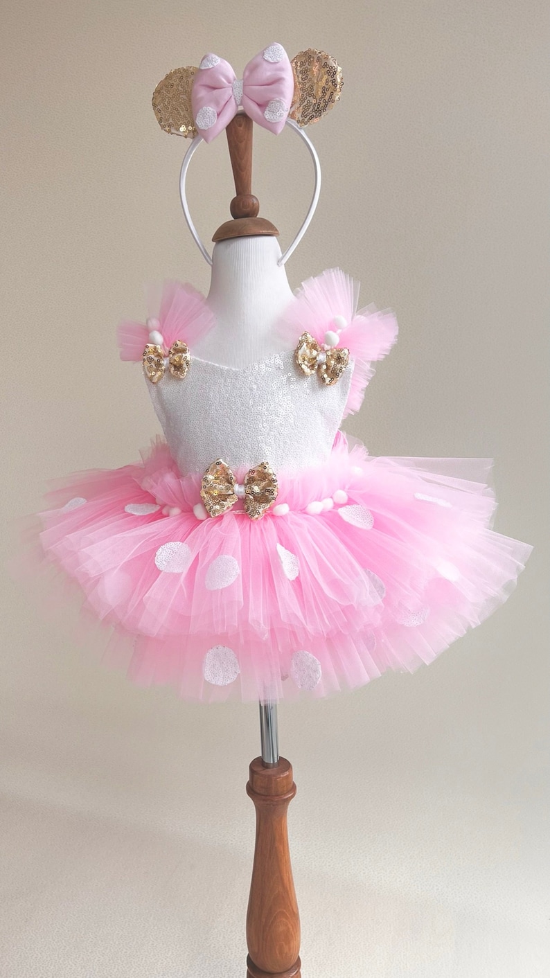 May include: A pink tulle tutu with white polka dots and a white bodice with gold bows. The tutu is worn by a mannequin and has a gold headband with pink bow and gold ears.