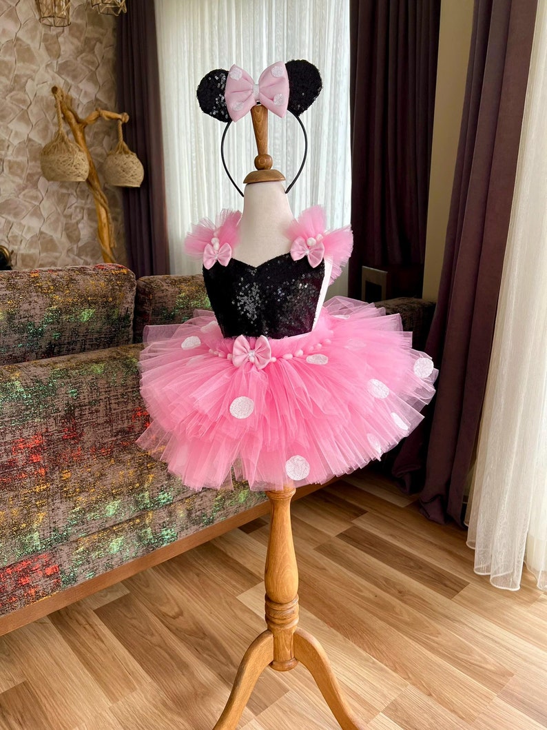 Pu&ograve; includere: Tutu rosa e nero con pois bianchi e un corpetto nero di paillettes. Il tutu &egrave; su un manichino di legno e ha un cerchietto nero con orecchie rosa e un fiocco.