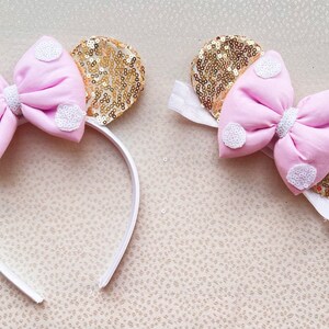 May include: A pink bow with white polka dots on a gold sequin headband. The headband is white and has a white bow with gold sequins.