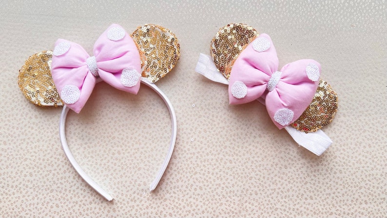 May include: A pink bow with white polka dots on a gold sequin headband. The headband is white and has a white bow with gold sequins.