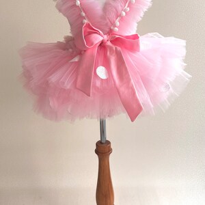 May include: A pink tulle tutu dress with white polka dots and a pink satin bow. The dress has a ruffled neckline and white pom-poms around the neckline.
