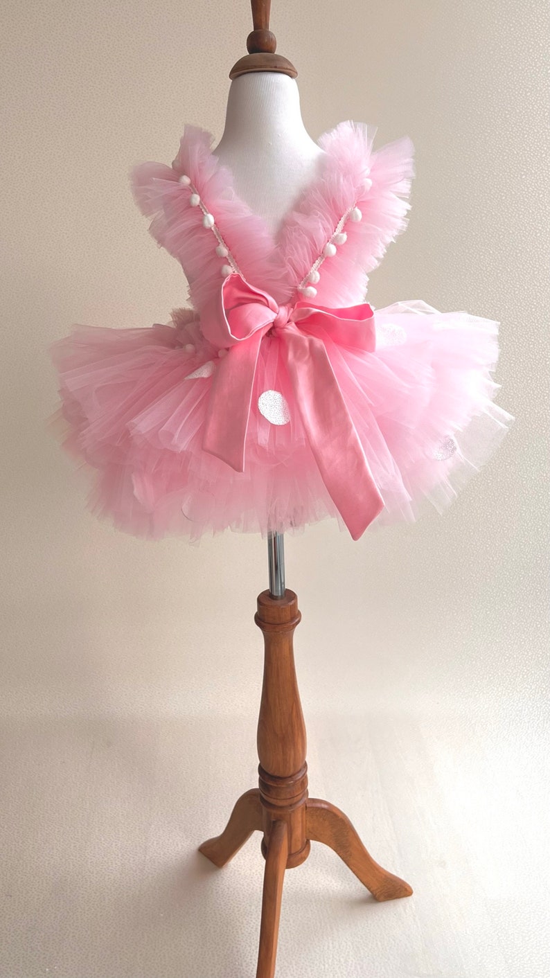 May include: A pink tulle tutu dress with white polka dots and a pink satin bow. The dress has a ruffled neckline and white pom-poms around the neckline.