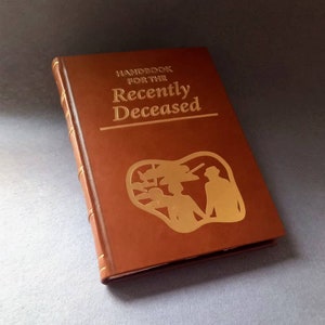 May include: A brown hardback book titled "Handbook for the Recently Deceased" with gold lettering and a gold embossed illustration. The book has gold page edges and a decorative design on the cover.