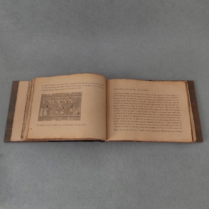 K&ouml;nnte beinhalten: Ein offenes Buch mit gealterten, vergilbten Seiten und einem dunklen Holzdeckel. Die linke Seite zeigt eine Schwarz-Wei&szlig;-Illustration von Figuren, w&auml;hrend die rechte Seite mit gedrucktem Text gef&uuml;llt ist. Das Buch ist auf einer grauen Oberfl&auml;che ge&ouml;ffnet.