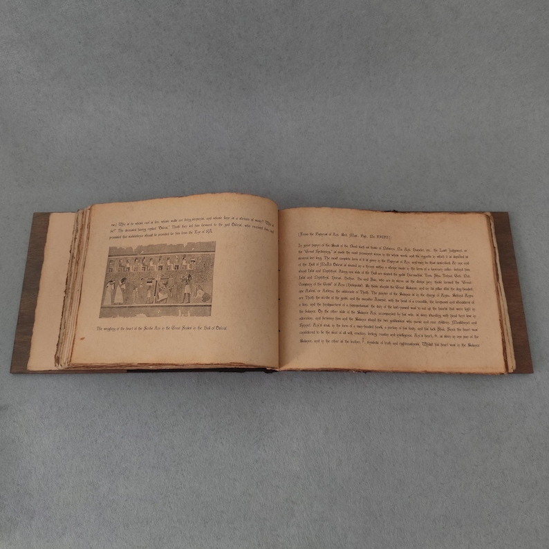K&ouml;nnte beinhalten: Ein offenes Buch mit gealterten, vergilbten Seiten und einem dunklen Holzdeckel. Die linke Seite zeigt eine Schwarz-Wei&szlig;-Illustration von Figuren, w&auml;hrend die rechte Seite mit gedrucktem Text gef&uuml;llt ist. Das Buch ist auf einer grauen Oberfl&auml;che ge&ouml;ffnet.