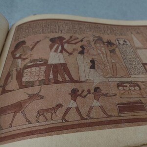 K&ouml;nnte beinhalten: Eine Buchseite mit einer detaillierten Illustration einer alt&auml;gyptischen Szene. Das Kunstwerk zeigt Figuren in traditioneller Kleidung, Tiere und Hieroglyphen in Braun-, Rot- und Wei&szlig;t&ouml;nen. Das Bild vermittelt ein Gef&uuml;hl von Geschichte und kulturellem Erbe.