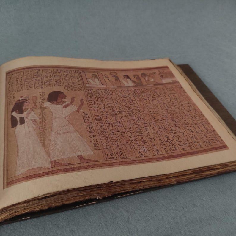K&ouml;nnte beinhalten: Ein offenes Buch mit gealterten, strukturierten Seiten. Die rechte Seite ist mit Hieroglyphen gef&uuml;llt, w&auml;hrend die linke eine gemalte Illustration von zwei Figuren in wei&szlig;en Roben zeigt. Die R&auml;nder des Buches sind abgenutzt, was auf Alter und Nutzung hindeutet.