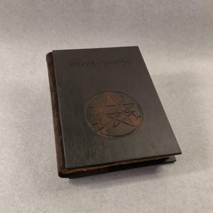 May include: A black leather-bound journal with a five-pointed star symbol embossed on the front cover. The journal has a brown leather spine and a title on the front cover that reads "The Book of Shadows".