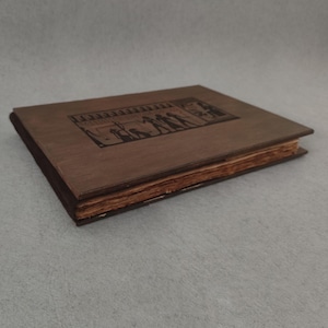 K&ouml;nnte beinhalten: Ein geschlossenes, antikes Buch mit einem braunen Holzdeckel und einem dunkelbraunen R&uuml;cken. Der Einband zeigt ein rechteckiges, graviertes Design mit Figuren. Die Seiten sind gealtert und haben einen gelblichen Farbton. Das Buch liegt auf einer grauen Oberfl&auml;che.