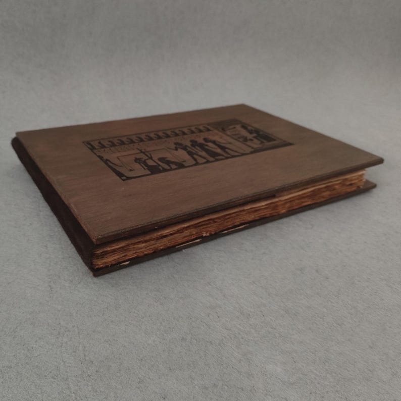 K&ouml;nnte beinhalten: Ein geschlossenes, antikes Buch mit einem braunen Holzdeckel und einem dunkelbraunen R&uuml;cken. Der Einband zeigt ein rechteckiges, graviertes Design mit Figuren. Die Seiten sind gealtert und haben einen gelblichen Farbton. Das Buch liegt auf einer grauen Oberfl&auml;che.