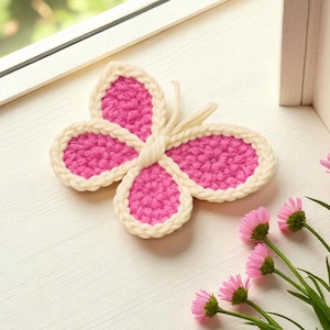 May include: A handmade crochet butterfly in a vibrant pink colour with a cream-coloured border. The butterfly is placed on a white surface near a window, with pink flowers in the foreground. The wings are detailed with a textured stitch.