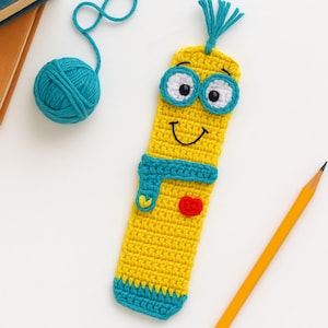 May include: A handmade yellow crochet bookmark with a smiling face, blue scarf, and red heart. The bookmark has blue eyes and a blue tassel. A ball of blue yarn and a yellow pencil are also in the image.