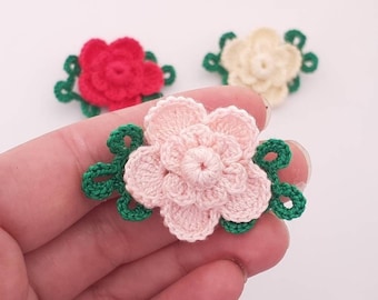 Puede incluir: Tres flores de ganchillo con hojas verdes. Las flores son rosas, rojas y crema.