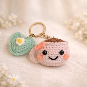 May include: A crocheted keychain featuring a smiling pink coffee cup with a brown coffee filling and a light green heart with a white flower. The keychain has a gold-colored ring.