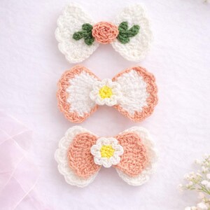 May include: Three handmade crochet hair bows. The bows are white, peach, and green, with floral accents. The top bow has a peach rose and green leaves. The middle and bottom bows have a white daisy in the centre.