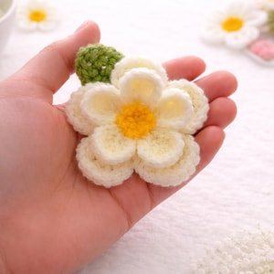 May include: A handmade crochet flower with white petals and a yellow center, accompanied by a green leaf. The flower is held in a hand, showcasing its delicate craftsmanship. This item is perfect for crafting and adding a touch of nature to any project.