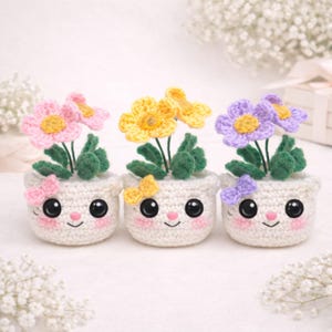 Puede incluir: Tres macetas de flores hechas a mano con ganchillo, cada una con una cara sonriente y un acento de mariposa. Las flores son rosas, amarillas y moradas, con hojas verdes. Las macetas son blancas y tienen un diseño lindo y de dibujos animados.