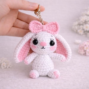 May include: A handmade white crochet bunny keychain with pink accents. The bunny has a pink bow, inner ears, and cheeks. It features black eyes and a gold-colored keychain ring. The bunny is approximately 4 inches tall.