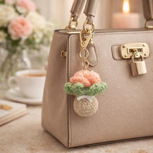Könnte beinhalten: Ein handgefertigter gehäkelter Pilz-Schlüsselanhänger in Pfirsich, Grün und Beige hängt an einer beigen Handtasche. Der Pilz hat einen pfirsichfarbenen Hut, grüne Lamellen und einen beigen Stiel. Der Schlüsselanhänger ist mit einem goldfarbenen Verschluss befestigt.