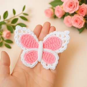 Op de afbeelding: Een gehaakte vlinder met roze vleugels en witte randen. De vlinder wordt in een hand gehouden, met roze rozen en groene bladeren op de achtergrond. De vleugels van de vlinder zijn zachtroze en de randen zijn wit omlijnd.