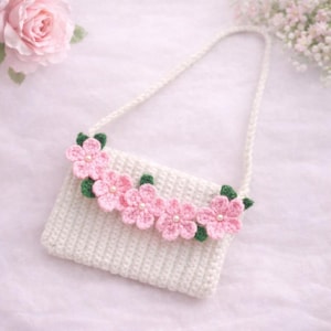 Pode incluir: Uma bolsa branca de crochê com uma alça, adornada com aplicações de flores rosa e folhas verdes. Cada flor tem um centro de pérola. A bolsa é um item feito à mão.