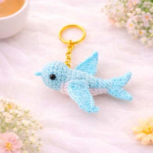Puede incluir: Un llavero de pájaro azul tejido a ganchillo con un anillo y una cadena dorados. El pájaro tiene ojos negros y detalles blancos en el pecho y el vientre. Las alas están extendidas y el pájaro está hecho de hilo suave.