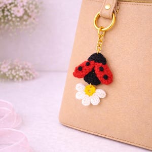 Könnte beinhalten: Ein handgefertigter Häkel-Schlüsselanhänger mit einem Marienkäfer mit roten Flügeln und schwarzen Punkten sowie einer weißen Gänseblümchen mit gelber Mitte. Der Schlüsselanhänger ist an einer goldfarbenen Kette und einem Ring befestigt, die an einer beigen Handtasche hängen.